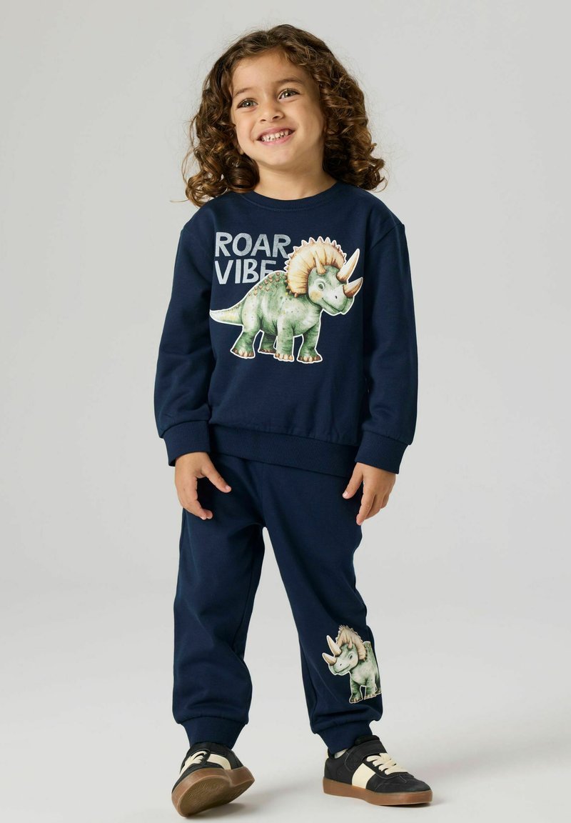 Smilende barn med krøllet hår iført navyblå sweatshirt og bukser med et grønt triceratops-dinosaurmotiv og teksten "ROAR VIBE".
