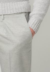 Grijze gebreide trui met geribbelde boorden, gecombineerd met lichtgrijze gestructureerde broek. Het zichtbare hand rust in de zak, waardoor het stofdetail zichtbaar is.