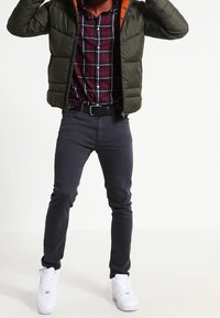 Uomo che indossa sneaker bianche, jeans scuri slim, una camicia a quadri rossi e blu, e un giubbotto imbottito verde aperto con fodera arancione.