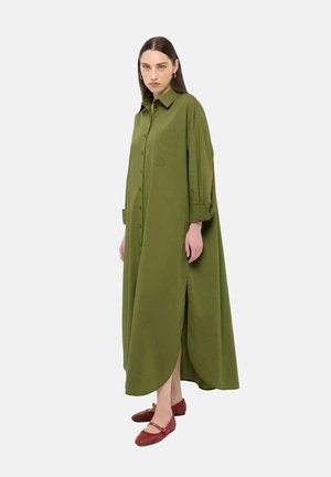 Vrouw draagt een lange olijfgroene jurk met knopen en opgerolde mouwen, en rode gevlochten Mary Jane flats, staand tegen een effen achtergrond.