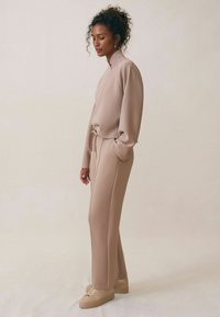 Beżowy zestaw odzieży domowej składający się z krótkiego topu z długim rękawem, o wysokiej szyi i regulowanym pasie, w zestawie z dopasowanymi spodniami o tapered kroju oraz beżowymi sneakersami.