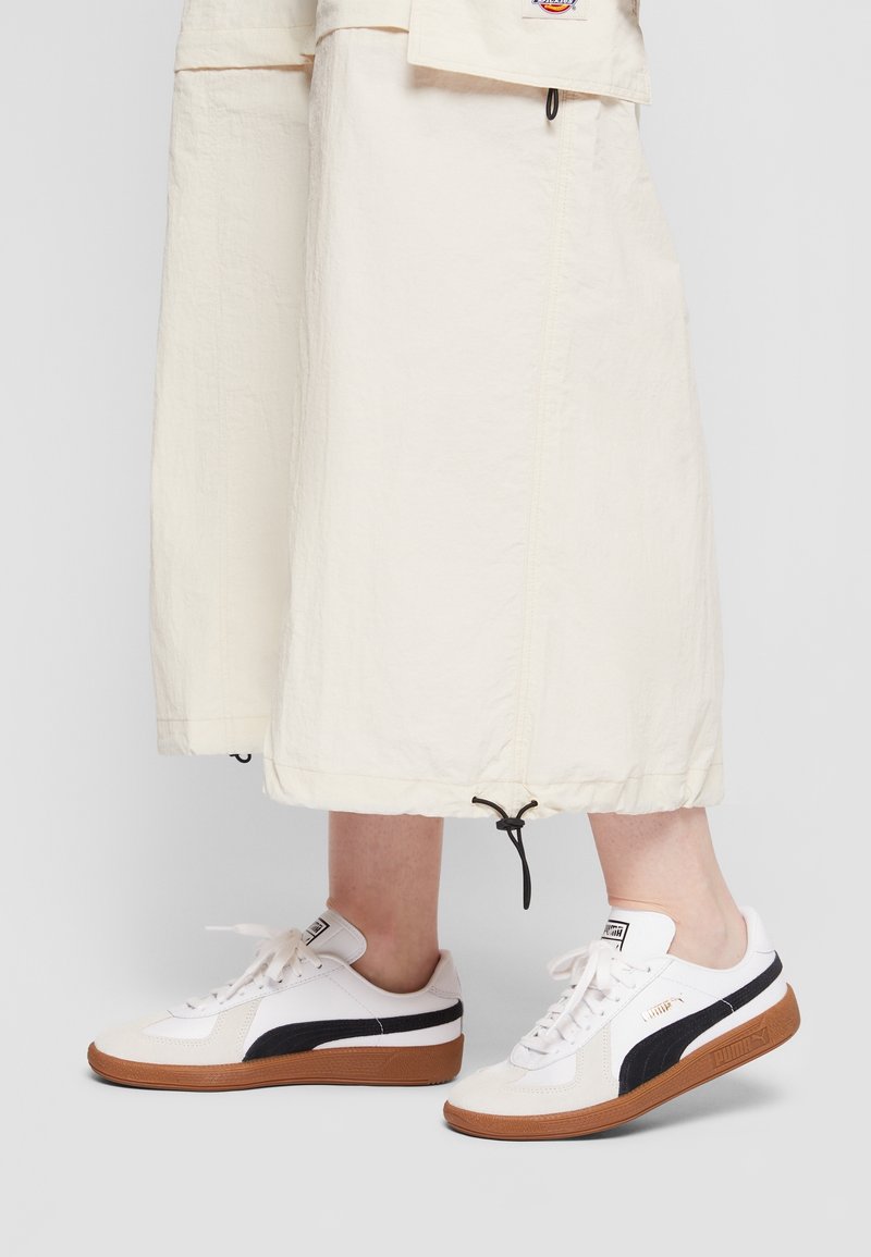 Jambes portant un pantalon crème large longueur cheville avec un cordon noir et des baskets blanches Puma avec des rayures noires et des semelles marron sur fond uni.