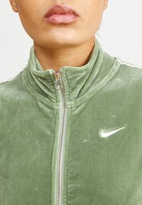 Chaqueta de felpa verde con cremallera, que presenta un cuello alto, cremallera plateada y un logo de Nike blanco en el pecho. Textura suave con un brillo sutil.