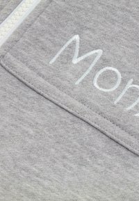 Grijs sweatshirtstof met een zachte textuur, voorzien van witte geborduurde tekst en een ritsluiting aan de voorkant. Eenvoudig ontwerp, geen patronen.