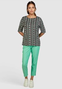 Schwarzes Shirt mit buntem geometrischem Muster, mit kurzen Ärmeln; kombiniert mit mintgrünen Hosen, schmaler Schnitt, und weißen Schuhen.
