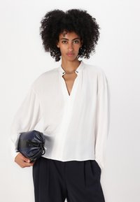 Blusa bianca a maniche lunghe con scollo a V profondo e leggere arricciature sulle spalle. Il tessuto appare leggero e liscio. Tiene una pochette blu navy.
