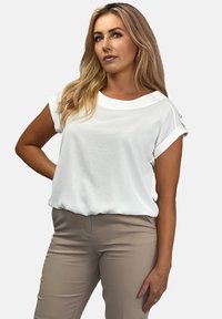 Biała bluzka krótki rękaw o luźnym kroju z podwiniętymi rękawami. Gładka tekstura, design z okrągłym dekoltem, noszona z beżowymi dopasowanymi spodniami.