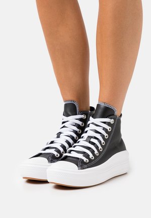 Benedenbenen met zwarte hoge sneakers met witte veters en dikke witte zolen op een lichte achtergrond.