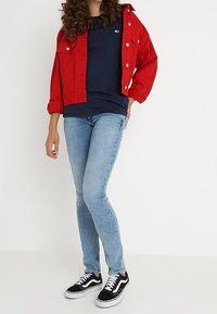 Chaqueta de mezclilla roja con botones plateados, camiseta azul marino, jeans ajustados de color azul claro y zapatillas negras con detalles en blanco. Conjunto casual.