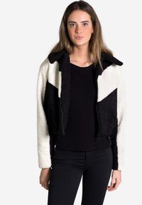 Chaqueta recortada de pelusa con bloques de color en blanco y negro, con cierre de cremallera y cuello. Lleva encima un suéter negro de canalé.