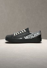 Baskets basses noires avec lacets blancs et texte blanc en gras "MOSCHINO" sur le côté, présentées sur une surface et un fond neutres.