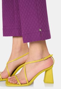 Pantalons violets à motifs avec une finition texturée, dotés d'un bouton décoratif à l'ourlet. Associés à des sandales à talons hauts jaunes à lanières.