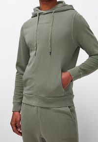 Felpa verde con cappuccio in morbido misto cotone, caratterizzata da lacci, tasca a marsupio e logo testuale sul petto. Design semplice e aderente.