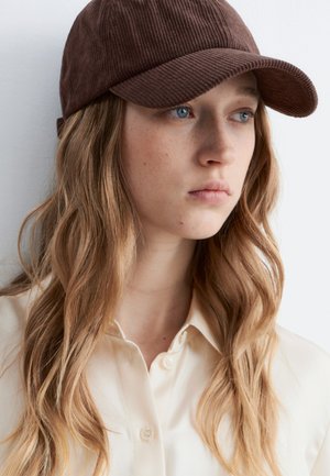 Jeune femme aux longs cheveux blonds ondulés, portant une casquette en velours côtelé marron et une chemise crème boutonnée, regardant sur le côté.