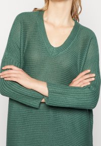 Grön stickad tröja med V-hals, ribbat mönster, långa ärmar och sprund i fållen, med en lös passform.