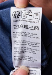 Skötselråd på tyg. Vit med svart text, som anger materialdetaljer: 95% bomull, 5% elastan, tvättinstruktioner och symboler.