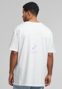 Bela bombažna majica z ohlapnim krojem. Na hrbtni strani je tiskan vijoličen motiv perja in napis. Kratki rokavi in okrogli izrez.