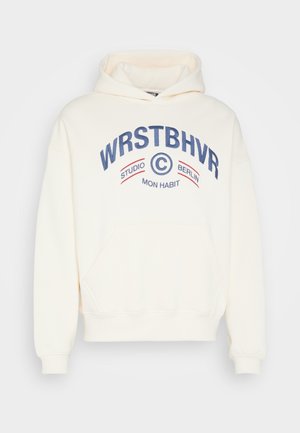 Bluza z kapturem w kolorze kremowym z kieszenią z przodu, niebiesko-czerwony napis "WRSTBHVR Studio Berlin Mon Habit" na piersi.