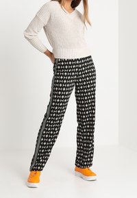 Ljusbeige stickad tröja med V-ringning; mönstrade svarta byxor med geometriska former, gröna sidopaneler och knallorange sneakers.