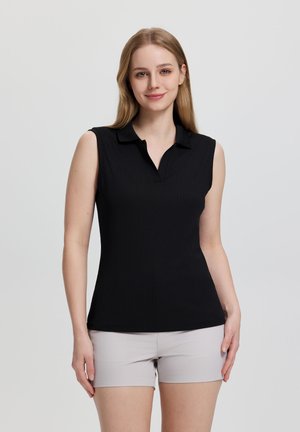 Mujer joven con cabello rubio largo, vistiendo una camisa polo negra sin mangas y pantalones cortos beige claro, de pie frente a un fondo liso.