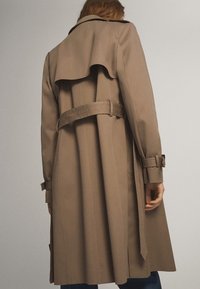 Personne portant un long trench-coat beige avec une ceinture et des poignets bouclés, vue de dos sur un fond clair uni.