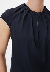 Navyblauwe blouse met korte puffmouwen, geplooide halslijn en een sleutelgatopening aan de voorkant. Glad stof met een zachte textuur.