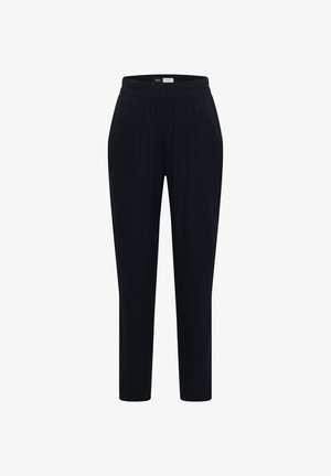 Femme souriante portant un haut beige à manches courtes, un pantalon bleu marine et des baskets beige, debout devant un fond beige uni.