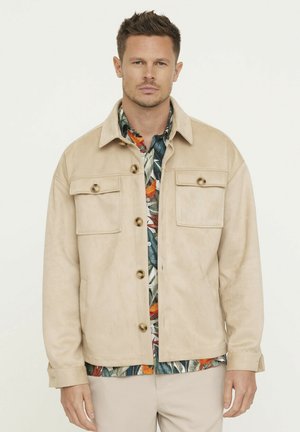Veste en suède beige avec deux poches avant, grands boutons marron et un col, portée sur une chemise à motif tropical. Texture douce, design décontracté.