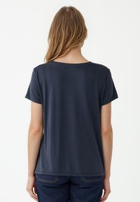 Granatowa bluzka z krótkim rękawem i okrągłym dekoltem, wykonana z gładkiego materiału. Widok z tyłu pokazuje luźny krój i lekko zaokrąglony dół.