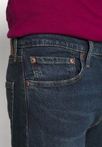 Mörkblå denimjeans med kopparnitar, rak passform och synliga sömnadsdetaljer; delvis bild som visar en vinröd skjorta ovanför.