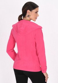 Felpa in pile di un vivace colore rosa, con un ampio cappuccio arrotondato, maniche lunghe e una texture morbida. Indossata con pantaloni neri attillati.