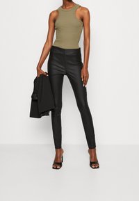 Pantaloni skinny neri in ecopelle con vita alta, abbinati a una canotta aderente color oliva. Il modello tiene un blazer nero e indossa sandali con la punta aperta.