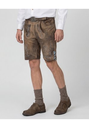 Mann trägt traditionelle braune Lederhose mit Stickerei, braune gestrickte Socken, braune Schuhe und ein weißes Langarmhemd, stehend.