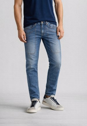 Uomo che indossa jeans blu sbiaditi con grandi tasche frontali a toppa, scarpe da ginnastica bianche con lacci neri e una maglietta blu navy.