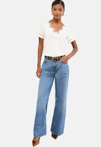 Donna che indossa una blusa bianca a maniche corte con scollo a V in pizzo, jeans blu a vita alta con gamba larga, cintura marrone e scarpe a punta marroni.