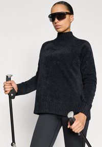 Melns, teksturēts džemperis ar apkakli, kopā ar melnām legingiem. Modelis tur soļu nūjas un valkā tumšas, lielizmēra saulesbrilles.