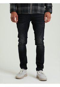 Jeans neri slim-fit con leggero sbiadimento, abbinati a sneakers di colore chiaro. Il modello indossa una camicia a quadri sopra una maglietta grigia.