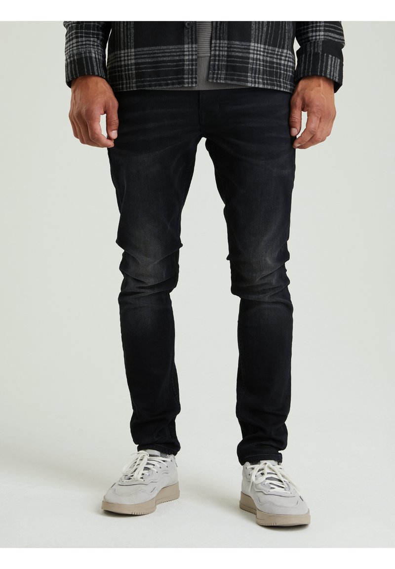 Jeans neri slim-fit con leggero sbiadimento, abbinati a sneakers di colore chiaro. Il modello indossa una camicia a quadri sopra una maglietta grigia.