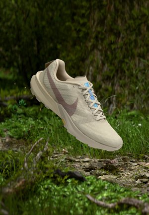 Lyst beige løbesko med et brunt swoosh-logo, svævende over en grøn skovbund med små planter og en stenet sti.
