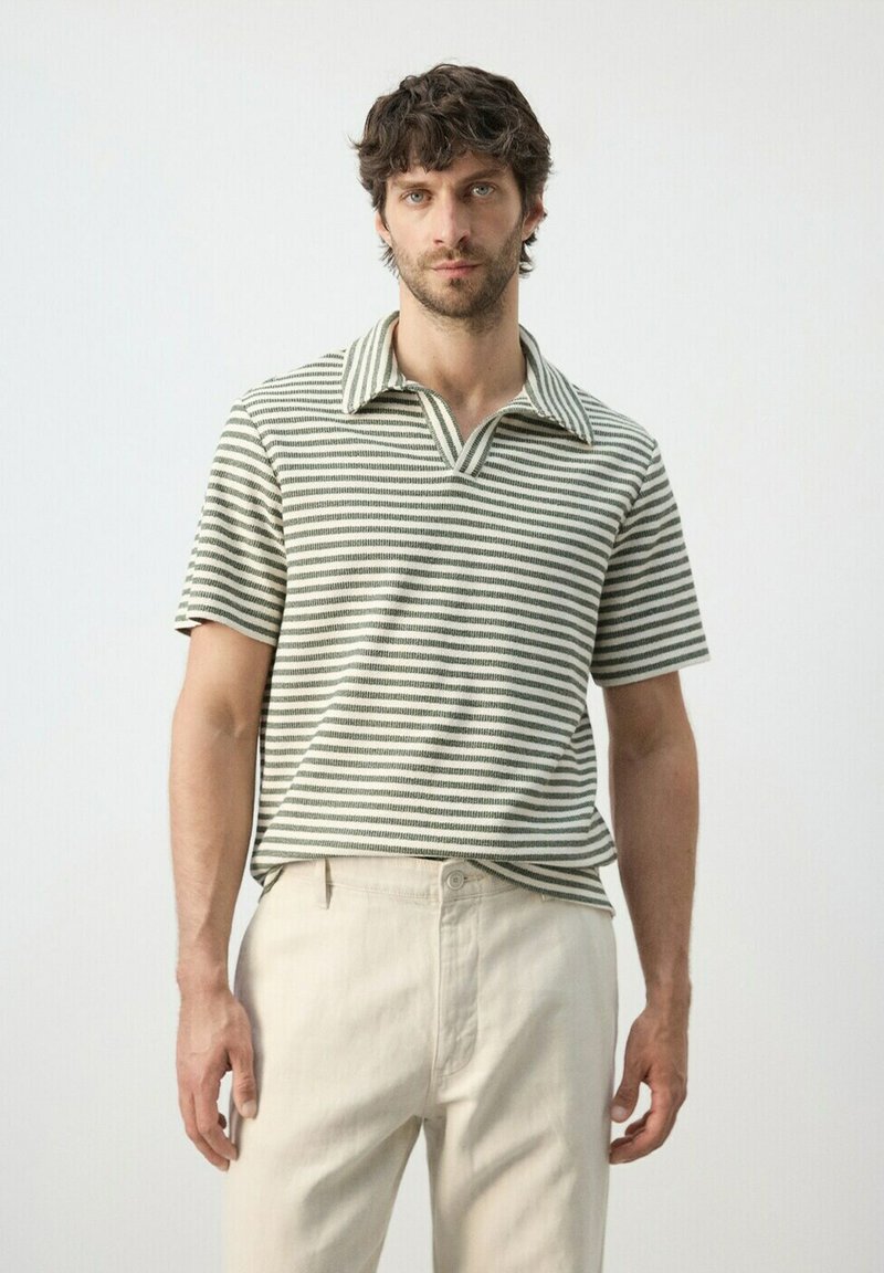 Homme aux cheveux bouclés et foncés, portant une chemise à col rayée à manches courtes et un pantalon beige clair, debout devant un fond uni clair.