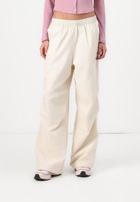 Vidje-byxor i ett ljusbeige tyg, med resårmidja och slät yta. Bärs med rosa sportskor.