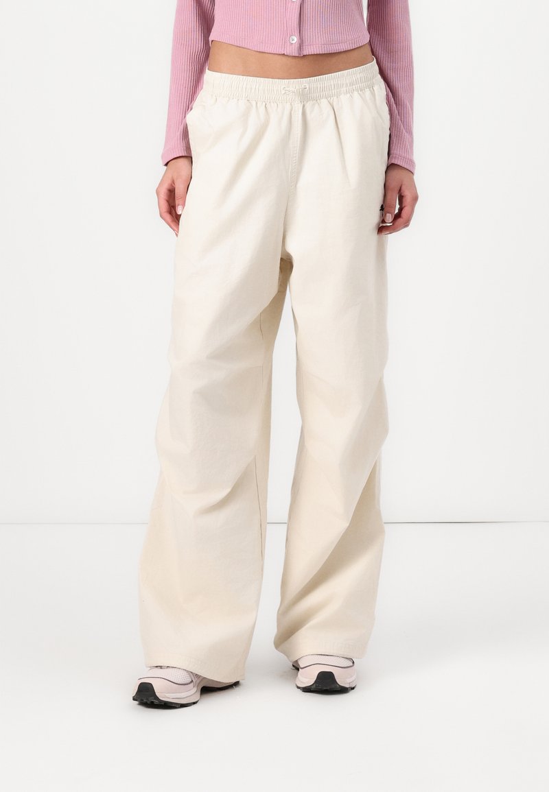 Vidje-byxor i ett ljusbeige tyg, med resårmidja och slät yta. Bärs med rosa sportskor.