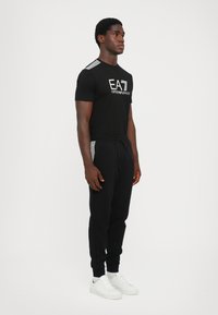 Svart t-skjorte med hvit "EA7 EMPORIO ARMANI" logo, kombinert med svarte joggebukser med hvite stripeakenter og hvite sneakers.