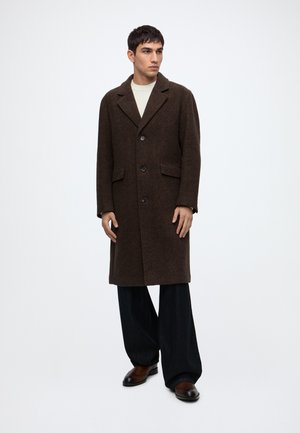 Homme debout portant un long manteau marron foncé boutonné, un pantalon noir à jambes larges et des chaussures marron foncé sur fond blanc uni.