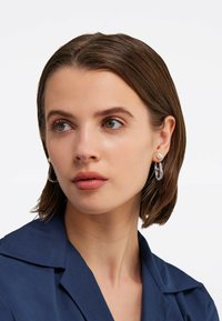Cerceii hoop argintii au un design cu linkuri transparente și un accent de perlă. Subiectul poartă un top cu guler de culoare albastră închis.