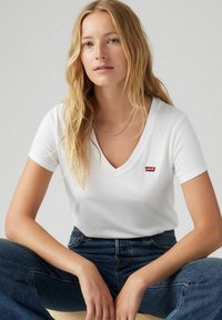 Camiseta blanca de cuello en V con un pequeño logo rojo de Levi's en el pecho izquierdo. La tela parece suave y el ajuste es casual, combinado con jeans azules.