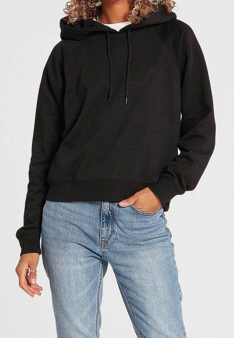 Personne aux cheveux bouclés portant un sweat à capuche noir et un jean bleu clair, une main dans la poche, sur un fond blanc uni.