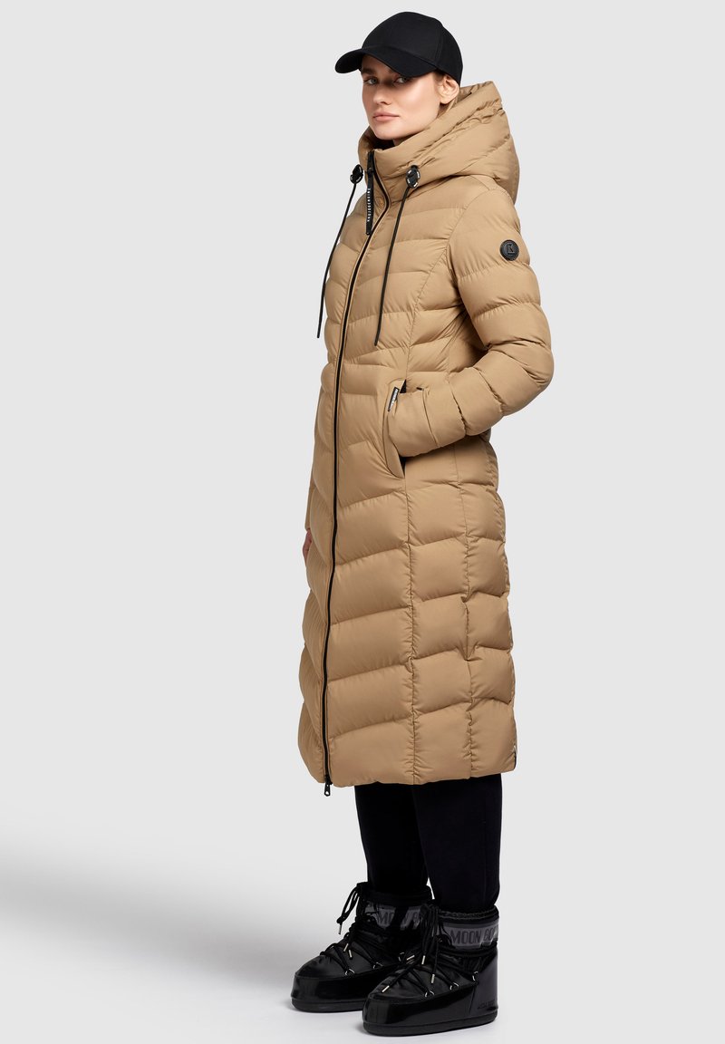 Langer, beiger Daunenmantel mit hohem Kragen, schwarzen Reißverschlüssen und Kordeln. Kombiniert mit schwarzem Schuhwerk mit glänzender Oberfläche.