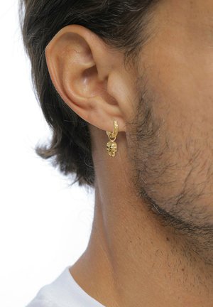 Nahaufnahme eines Ohrs einer Person, die einen goldenen Creolen-Ohrring mit einem detaillierten Totenkopf-Anhänger vor weißem Hintergrund trägt.