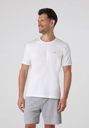 Uomo con capelli corti castani che indossa una camicia bianca a maniche corte e pantaloncini grigi, in piedi e sorridente mentre guarda a destra.